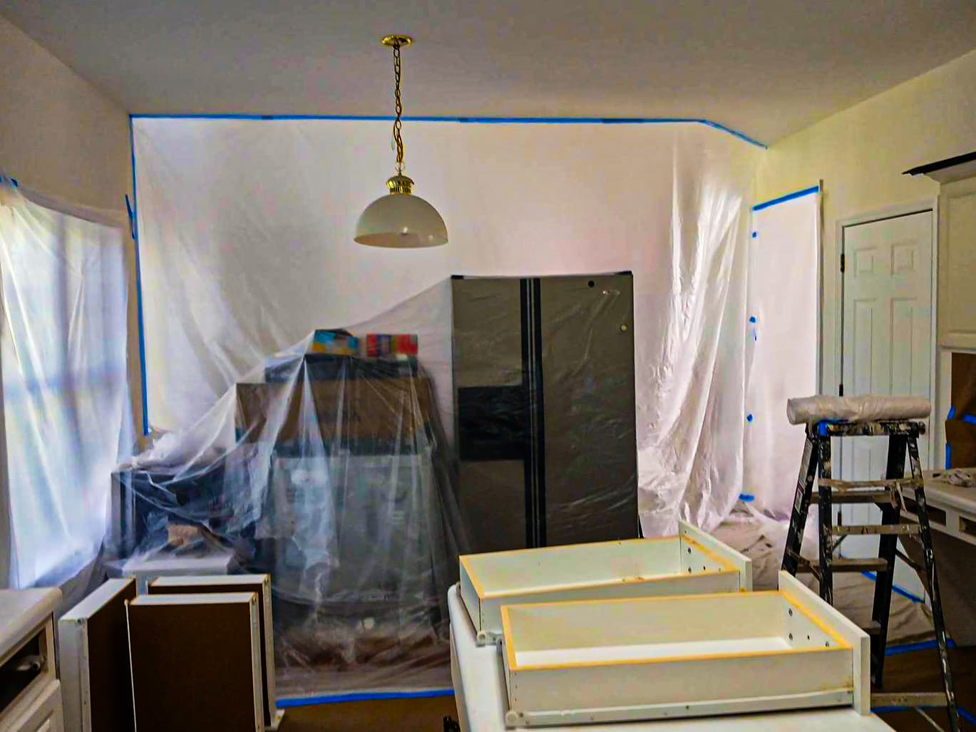 Interior painting project with furniture, cabinets, floors, walls, and fixtures protected with plastic, tape, and coverings.